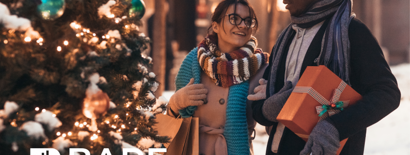 A man and woman holding presents and shopping bags standing beside a Christmas tree