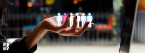 A close-up of a woman's hand with dark nail polish extended in front of a laptop screen, appearing to hold a row of digital human icons. The icons are semi-transparent, glowing white and pink, symbolizing data-driven decision-making and audience segmentation. The blurred background suggests an outdoor café or urban setting. A white logo with stylized text 'BR' is visible in the bottom left corner.