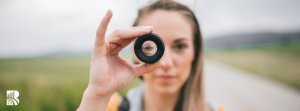 A young woman with long blonde hair holds a small camera lens up to her eye, looking through it. The background is blurred, showing an outdoor rural or suburban setting with a pathway and green fields. The lens creates a focused circular frame around her eye, emphasizing the concept of perspective and observation. A white logo with stylized text 'BR' is visible in the bottom left corner.