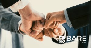 Close-up of professionals joining fists in a teamwork gesture, symbolizing ethics, collaboration, and social responsibility, with the BARE International logo.